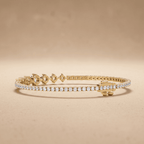 A gold tennis bracelet with a row of clear pavé-set stones and decorative gold clasp details, displayed on a warm beige background.