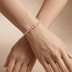 A delicate diamond tennis bracelet with round-cut center stones and smaller pave-set diamonds, shown worn on a model's wrist with crossed hands against a warm beige background.