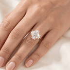 A close-up of a hand wearing a rose gold ring with a large clear round-cut center stone surrounded by a halo of smaller clear stones, resting on soft white fabric.
