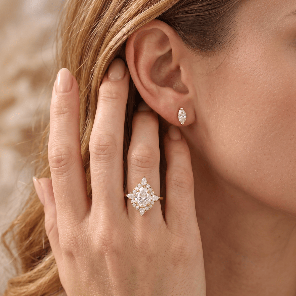 A close-up lifestyle shot of a woman wearing a gold ring with a diamond halo and a matching diamond stud earring. The ring features a marquise-cut center stone surrounded by a diamond cluster, set on a gold band.