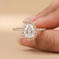A close-up of a hand holding a gold ring with a pear-cut clear center stone and small surrounding stones on a warm beige background.