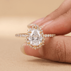 A close-up of a hand holding a gold ring with a pear-cut clear center stone and small surrounding stones on a warm beige background.