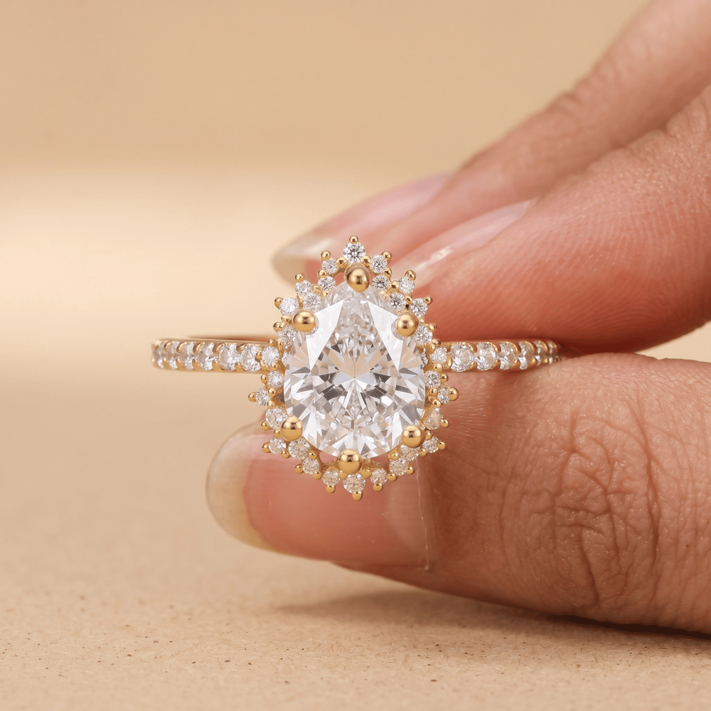 A close-up of a hand holding a gold ring with a pear-cut clear center stone and small surrounding stones on a warm beige background.