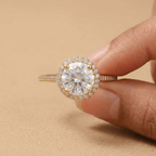 A close-up of a gold halo engagement ring with a large round clear center stone and smaller accent stones, held between fingers against a warm beige background. Per the color rules, clear gemstones do not contribute a color, so only the gold metal setting is counted.