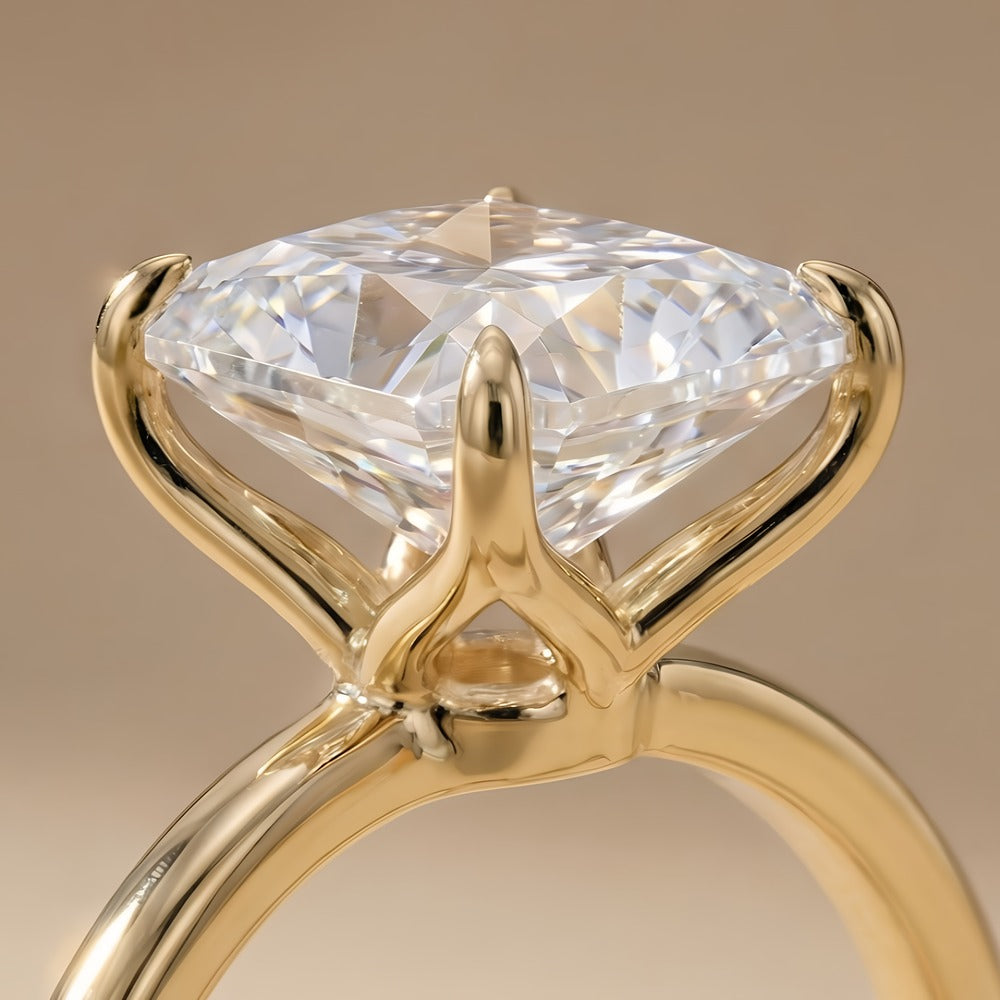 A close-up of a yellow gold solitaire ring featuring a large, clear round brilliant-cut diamond. The diamond is held in a four-prong setting on a polished band, set against a soft, warm beige background.