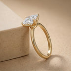 A gold ring with a large clear marquise-cut gemstone, resting on a textured beige stone block against a matching beige background.