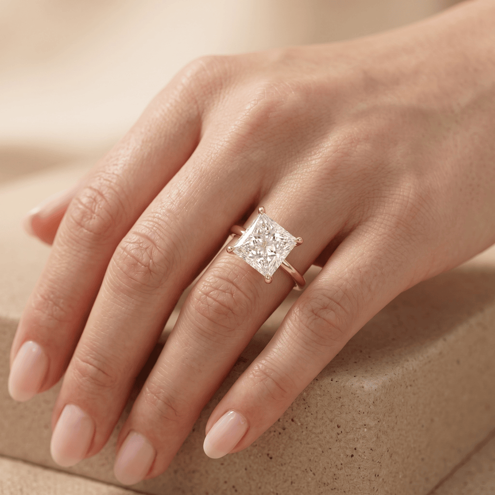 A close-up of a hand wearing a rose gold engagement ring with a princess-cut diamond. The ring is shown on the ring finger against a soft, warm beige background.