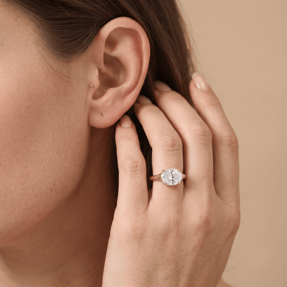 A close-up lifestyle shot of a woman wearing a rose gold ring with a round-cut clear gemstone on her finger, with her hand resting near her ear against a solid beige background.