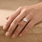 A close-up of a hand wearing a rose gold engagement ring with a large round brilliant-cut diamond. The hand is resting on a textured beige stone surface.
