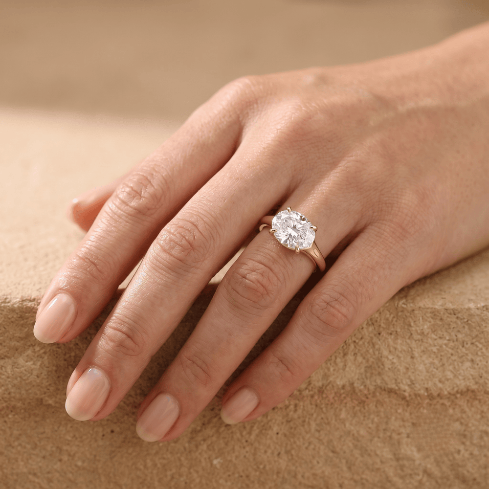 A close-up of a hand wearing a rose gold engagement ring with a large round brilliant-cut diamond. The hand is resting on a textured beige stone surface.