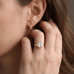 A close-up lifestyle shot of a woman wearing a rose gold ring with a round brilliant-cut diamond on her finger, with a matching rose gold hoop earring visible on her ear.
