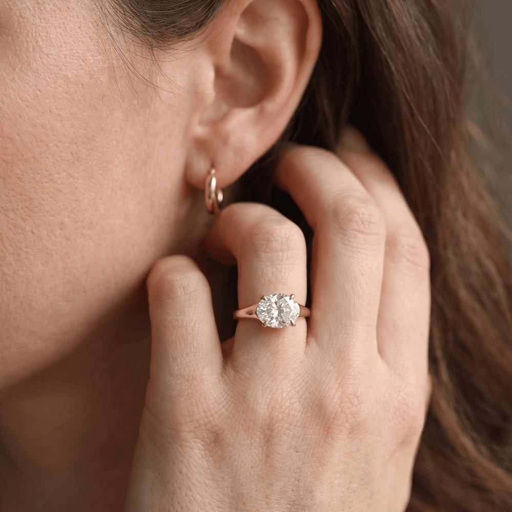 A close-up lifestyle shot of a woman wearing a rose gold ring with a round brilliant-cut diamond on her finger, with a matching rose gold hoop earring visible on her ear.