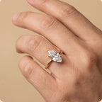 A close-up of a hand wearing a rose gold ring with a marquise-cut clear gemstone, set against a solid beige background.