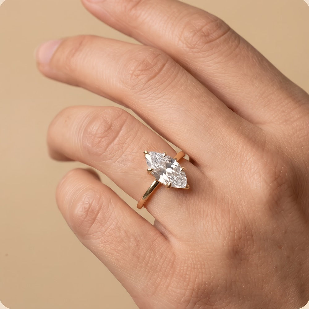 A close-up of a hand wearing a rose gold ring with a marquise-cut clear gemstone, set against a solid beige background.