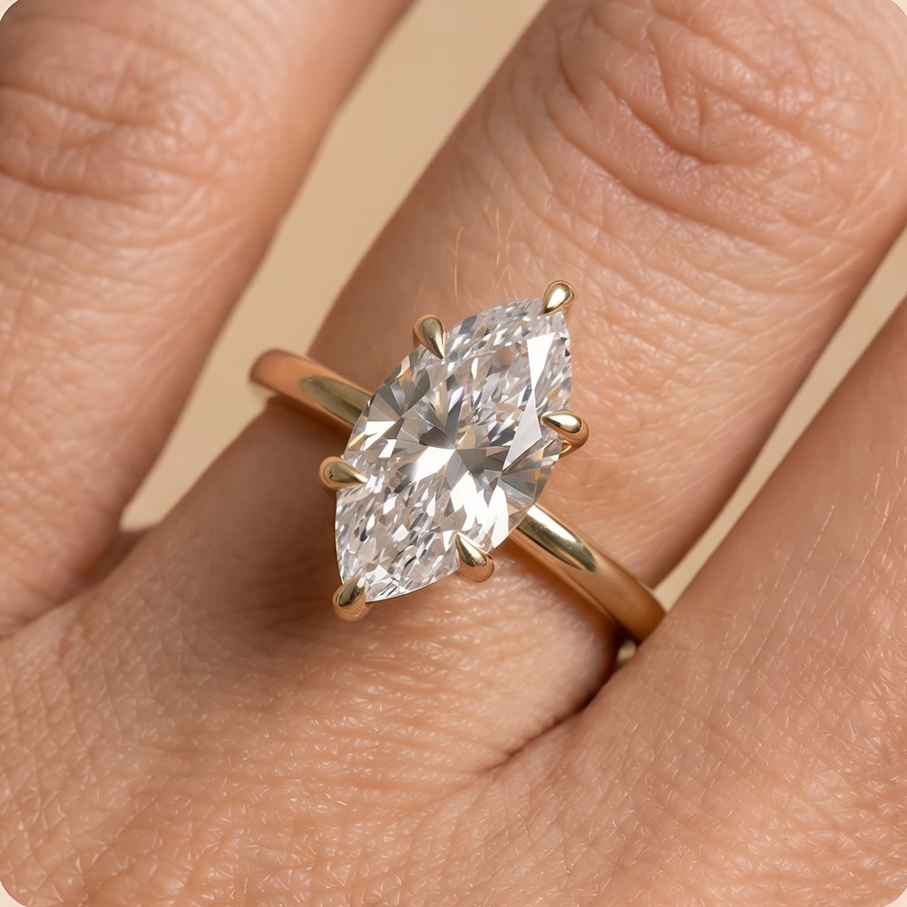 A close-up of a gold ring with a marquise-cut clear gemstone worn on a finger. The ring features a polished yellow gold band and prongs holding the stone, set against a soft beige skin background.