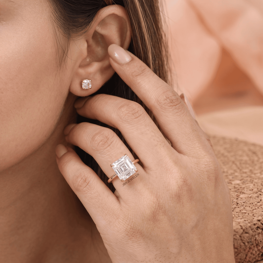 A close-up lifestyle shot of a woman wearing a rose gold ring with a large emerald-cut clear gemstone and a matching small stud earring.