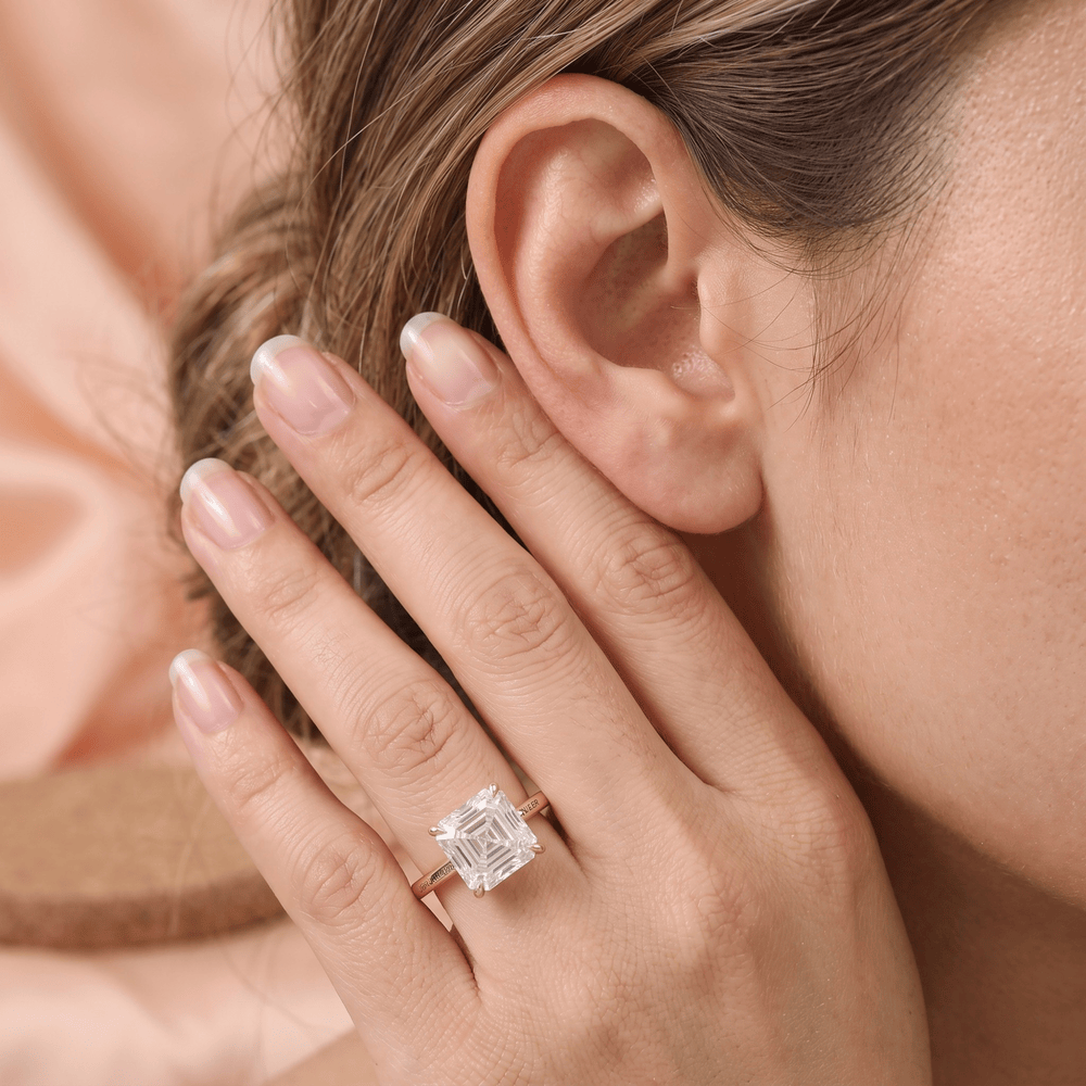 A close-up lifestyle shot of a woman wearing a rose gold ring with a square-cut clear gemstone on her finger, with her hand resting near her ear.