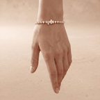 A delicate bracelet with a central cross charm and small geometric links, shown worn on a hand against a soft, warm beige background.