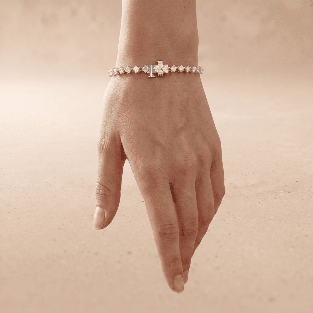 A delicate bracelet with a central cross charm and small geometric links, shown worn on a hand against a soft, warm beige background.