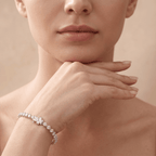 A close-up lifestyle shot of a model wearing a delicate diamond tennis bracelet on her wrist, with her hand resting near her chin against a soft beige background.