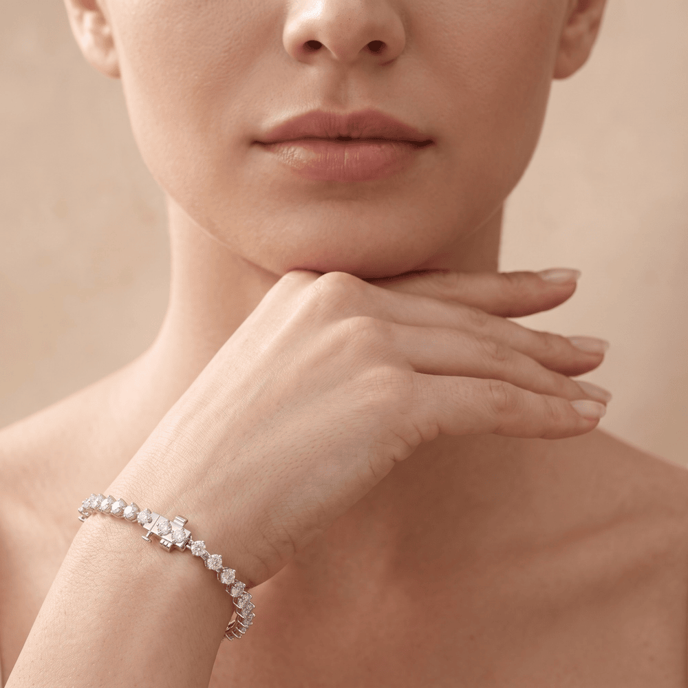 A close-up lifestyle shot of a model wearing a delicate diamond tennis bracelet on her wrist, with her hand resting near her chin against a soft beige background.