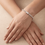 A close-up of a woman's hands and wrist wearing a delicate silver tennis bracelet with clear gemstones, resting on a textured beige surface.
