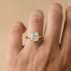 A close-up of a hand wearing a yellow gold ring with a large cushion-cut clear gemstone, set against a solid beige background.