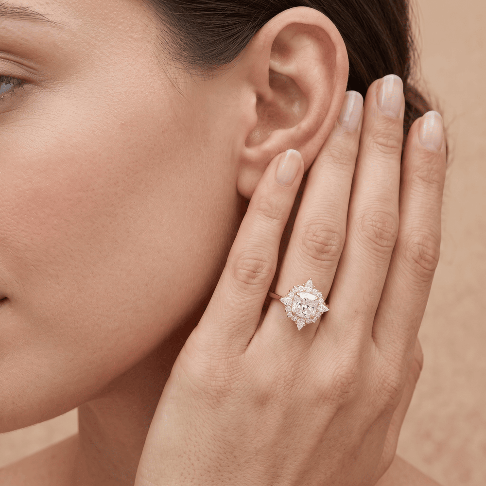 A close-up lifestyle shot of a woman wearing a diamond ring with a starburst cluster setting on her ring finger, with her hand resting against her ear.
