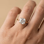 A close-up of a hand wearing a rose gold ring with a large cushion-cut clear gemstone. The ring is shown on the ring finger against a soft, solid beige background.