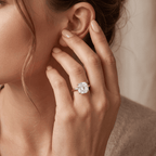 A close-up lifestyle shot of a woman wearing a silver-toned diamond engagement ring on her ring finger, with her hand resting near her ear against a soft beige background.