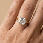 A close-up of a hand wearing a gold ring with a large clear cushion-cut gemstone, set against a soft beige background.