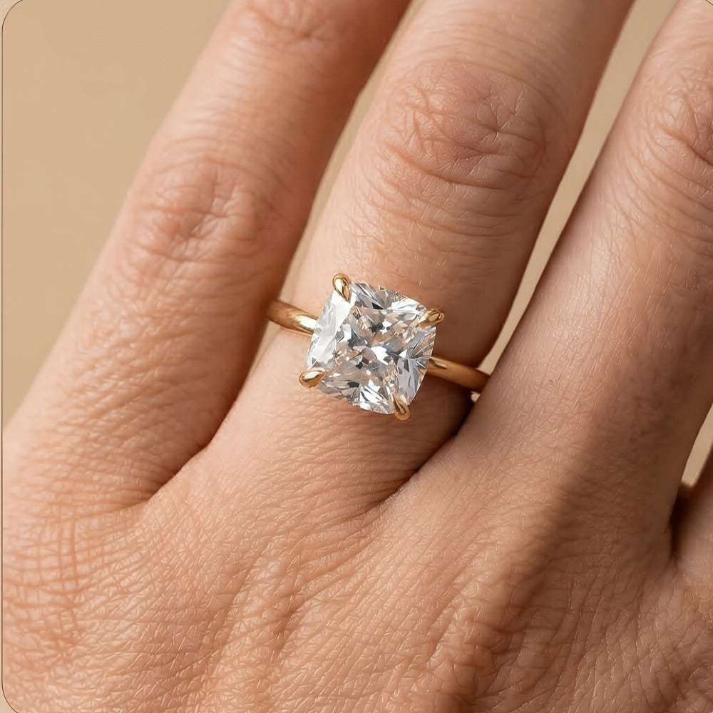 A close-up of a hand wearing a gold ring with a large clear cushion-cut gemstone, set against a soft beige background.