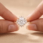 A close-up of a gold halo ring with a large round clear center stone and smaller surrounding stones, held between two fingers against a soft beige background.