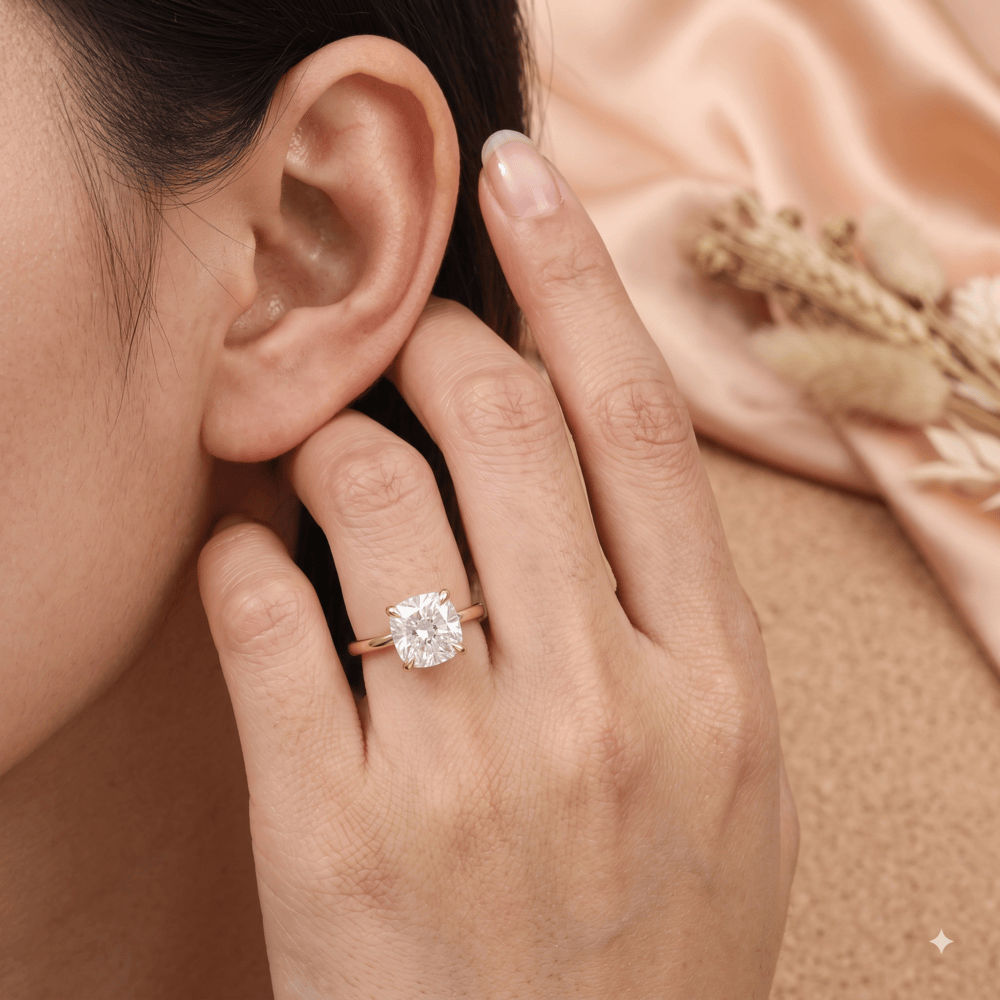 A close-up lifestyle shot of a woman's hand and ear wearing a rose gold ring with a cushion-cut clear gemstone, set against a soft peach satin background with dried flowers.