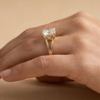 A close-up of a hand wearing a yellow gold ring with a large cushion-cut clear gemstone. The ring features a split shank design with small triangular gold details at the top of the stone.