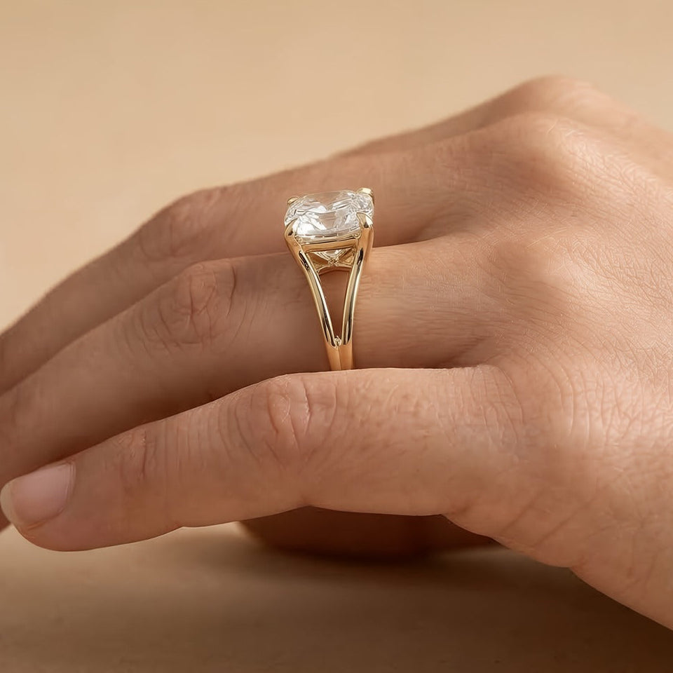 A close-up of a hand wearing a yellow gold ring with a large cushion-cut clear gemstone. The ring features a split shank design with small triangular gold details at the top of the stone.