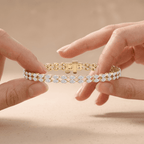 A close-up of a gold tennis bracelet with clear round-cut stones, held between two hands against a soft beige background.