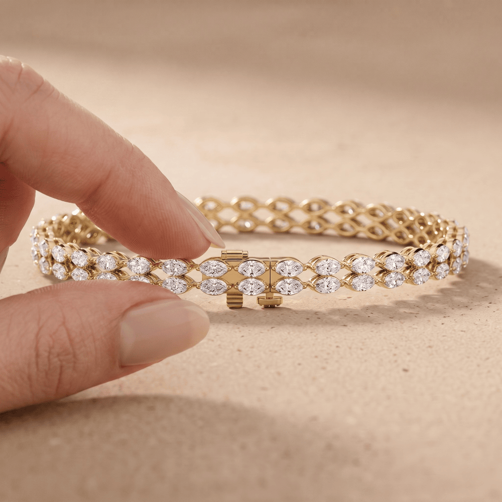 A close-up of a gold tennis bracelet with clear pavé stones, held gently by a person's fingers against a warm beige textured background.
