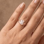 A close-up of a hand wearing a rose gold ring with an emerald-cut clear center stone and decorative side stones, set against a warm brown background.