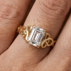 A gold ring with an emerald-cut clear center stone and decorative side stones, shown worn on a finger.