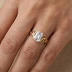 A gold ring with an emerald-cut clear center stone, worn on a finger. The ring features an intricate gold band with twisted details on either side of the stone.