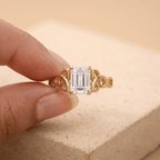 A gold ring with an emerald-cut clear center stone held between fingers against a warm beige background.