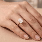 A close-up of a hand wearing a rose gold ring with a large rectangular emerald-cut clear gemstone, resting on a neutral beige surface.