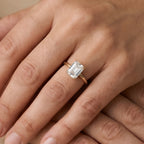 A close-up of a hand wearing a gold ring with an emerald-cut clear gemstone. The ring is shown on the ring finger against a soft, warm beige background.