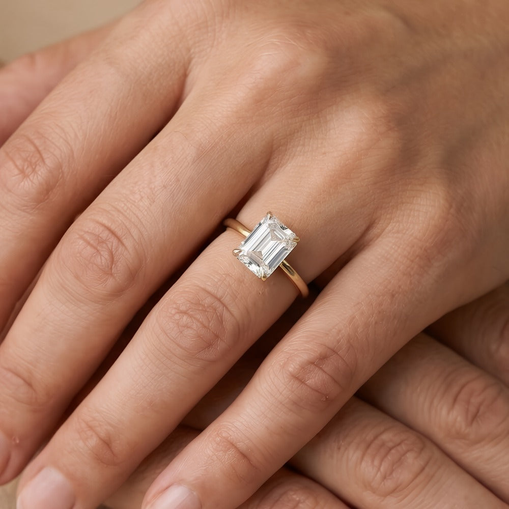 A close-up of a hand wearing a gold ring with an emerald-cut clear gemstone. The ring is shown on the ring finger against a soft, warm beige background.