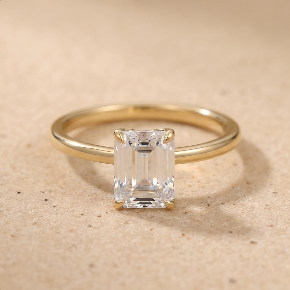 A yellow gold ring with a rectangular emerald-cut clear gemstone, photographed on a textured beige surface.