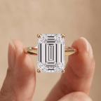 An emerald-cut diamond ring with a yellow gold band, held between two fingers against a soft beige background.