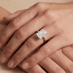 A close-up of a hand wearing an emerald-cut diamond ring on a silver band, resting on another hand against a neutral beige background.