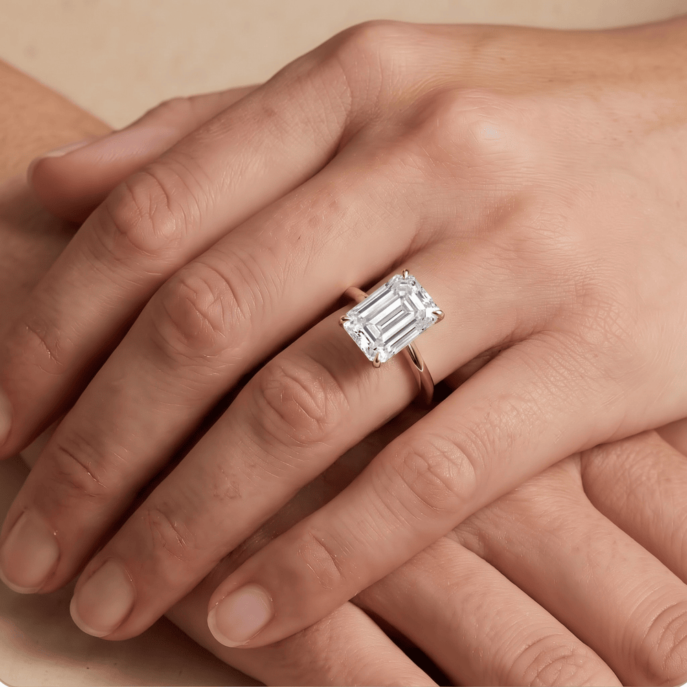 A close-up of a hand wearing an emerald-cut diamond ring on a silver band, resting on another hand against a neutral beige background.