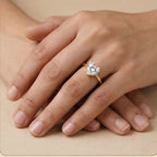 A heart-shaped diamond engagement ring on a gold band, shown worn on a person's hand resting on a beige surface.